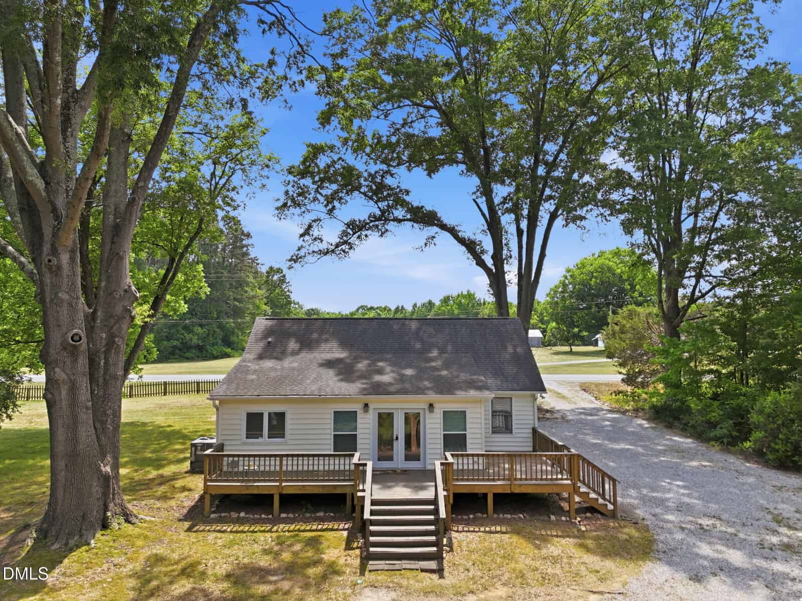 A small white house with a gray roof and large front porch, surrounded by tall trees and grassy areas, sits at the end of a gravel driveway on a sunny day.