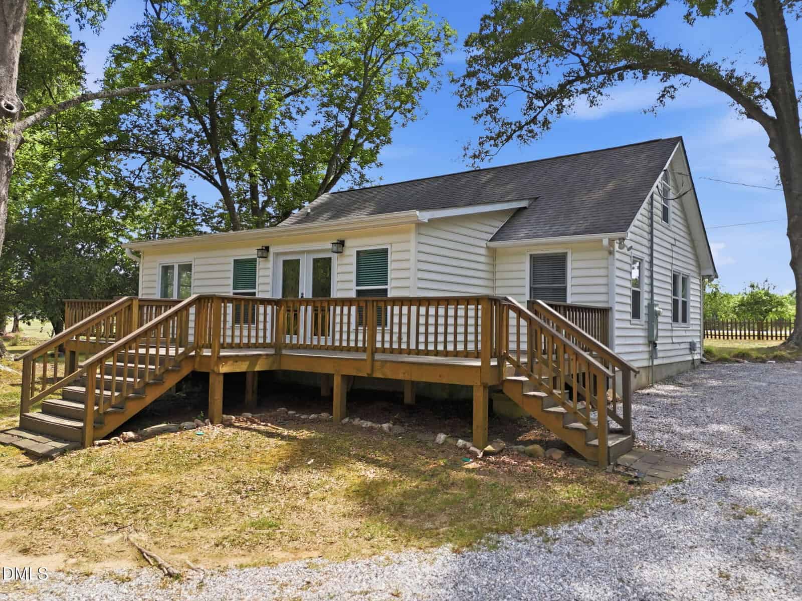 A small white house with green shutters and a black roof, featuring a large wooden deck and steps, surrounded by trees and a gravel driveway on a sunny day.