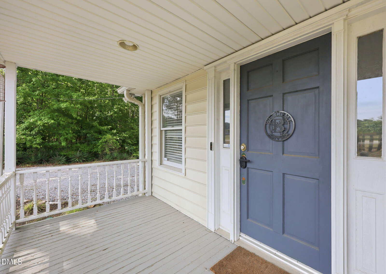 A covered porch with light-colored siding, a blue front door featuring a decorative emblem, white railings, and a doormat. Trees and gravel are visible in the background.