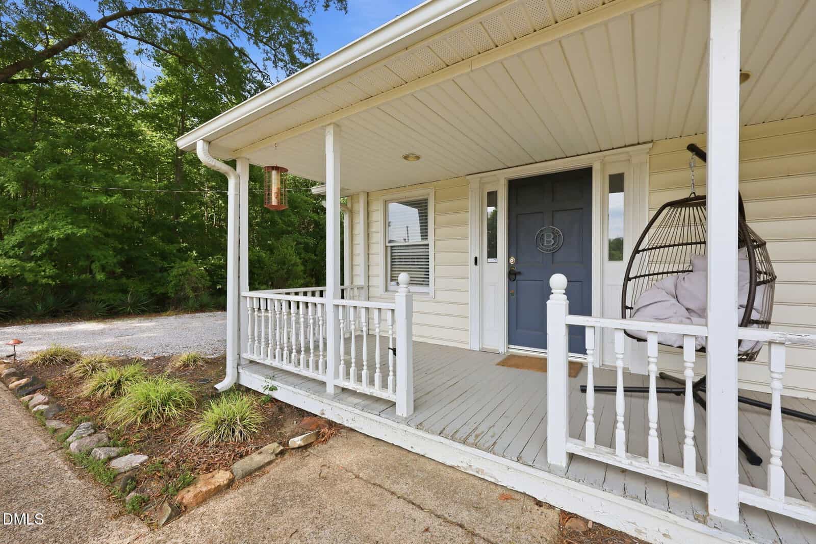 A white house porch with a blue front door, white railing, a hanging egg chair with cushions, and a small landscaped area beside a gravel driveway, surrounded by green trees.