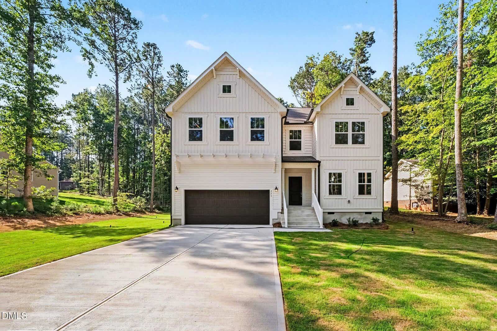 A modern two-story white house at 225 Rawhide Drive 1851, Louisburg, featuring a dark garage door, large windows, and a spacious driveway, is surrounded by green grass and tall trees under a blue sky.