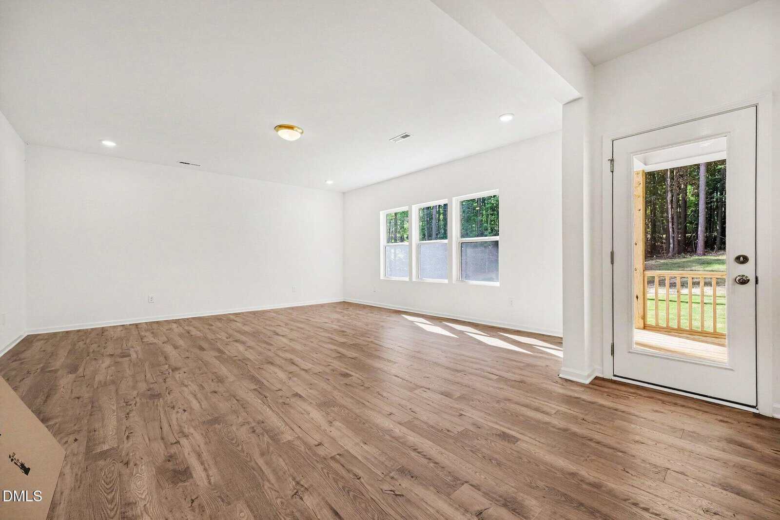 Bright empty room with wood flooring, white walls, three large windows, ceiling lights, and a glass door leading to a small wooden deck with a view of green grass and trees outside.