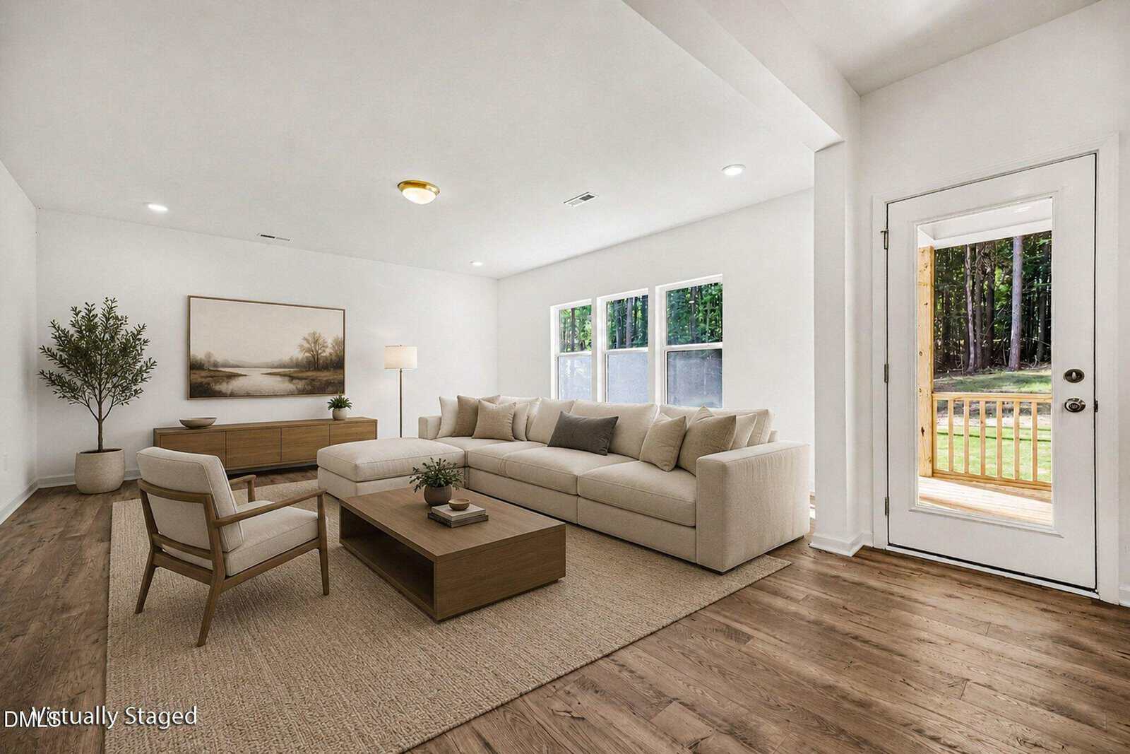 Bright living room with a large beige sectional, wooden coffee table, chair, sideboard, potted plant, floor lamp, large wall art, and a door leading outside. Three windows let in natural light.