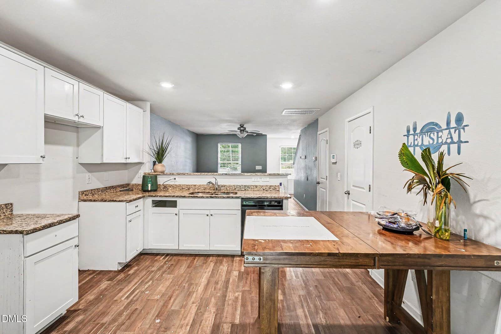 A modern kitchen with white cabinets, granite countertops, wooden floors, and a wooden dining table. The wall features a "Let's Eat" sign and there are plants for decoration, with natural light coming through windows.