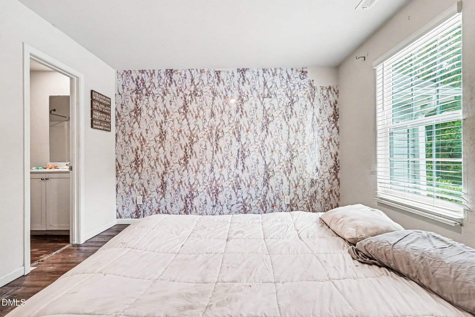 A minimalist bedroom with a mattress on the floor, neutral bedding, a marble-patterned accent wall, large window with blinds, and an open door leading to a bathroom.