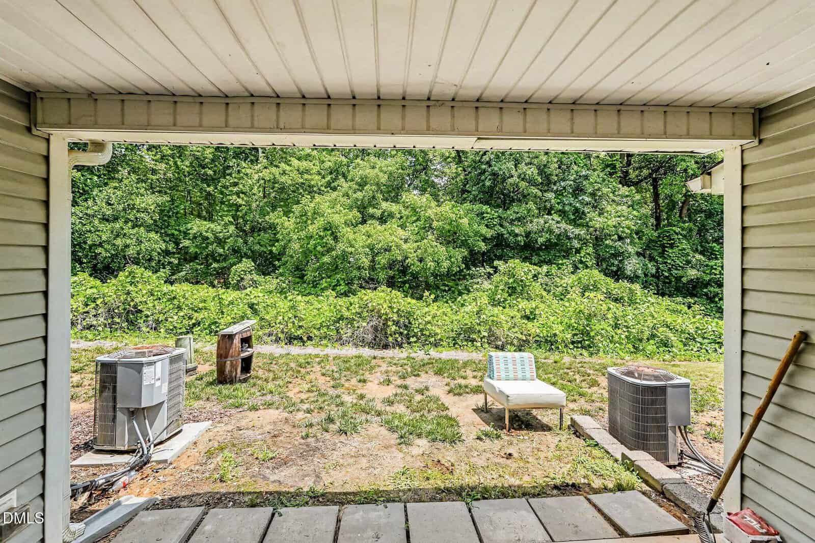 View from a covered patio showing two air conditioning units, a weathered grill, a lounge chair, and a grassy yard bordered by dense green trees in the background.