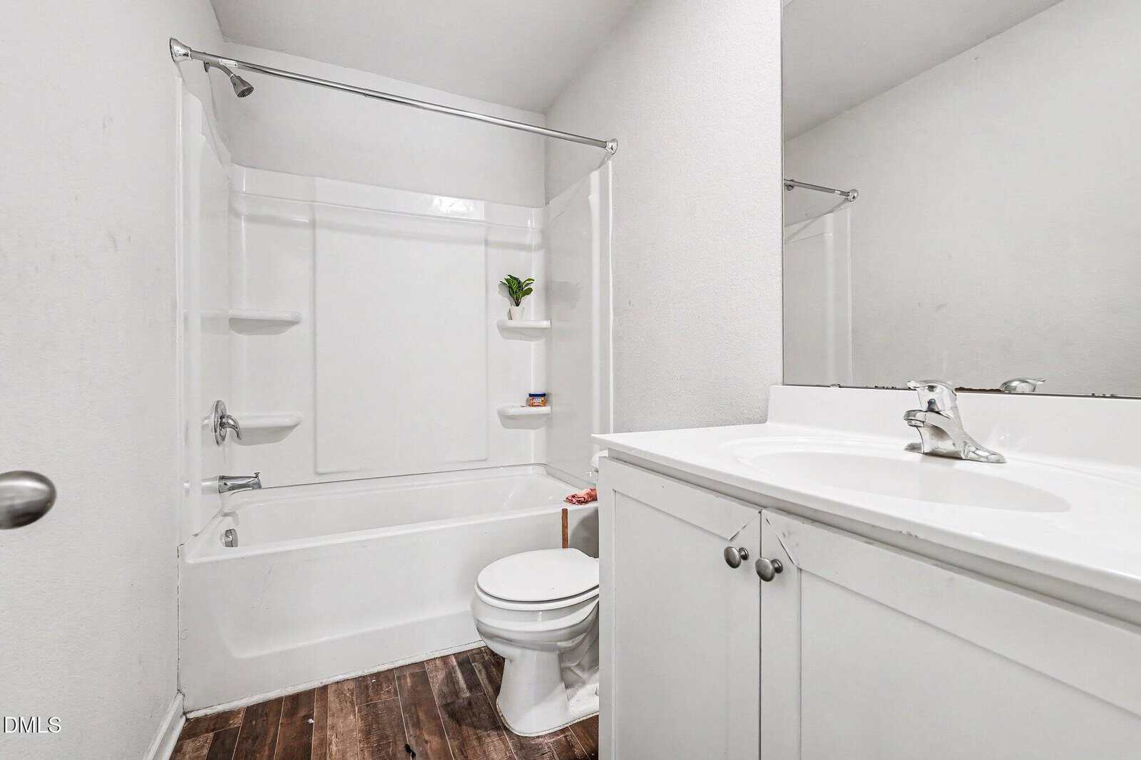 A modern bathroom with white walls, a white bathtub and shower, a toilet, a white vanity with a sink, a large mirror, and wood-look flooring; a small plant sits on a bathtub shelf.