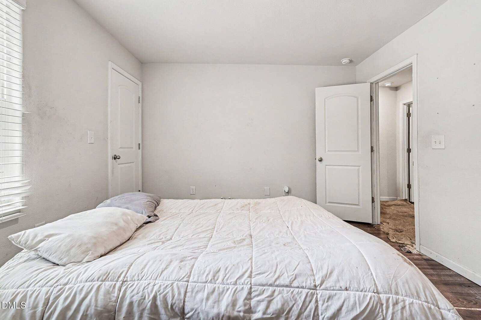 A simple bedroom with a bed against a wall, covered with a white comforter and two pillows. The room has white walls, an open door, and light coming through closed blinds on the left. The floor appears to be wooden.