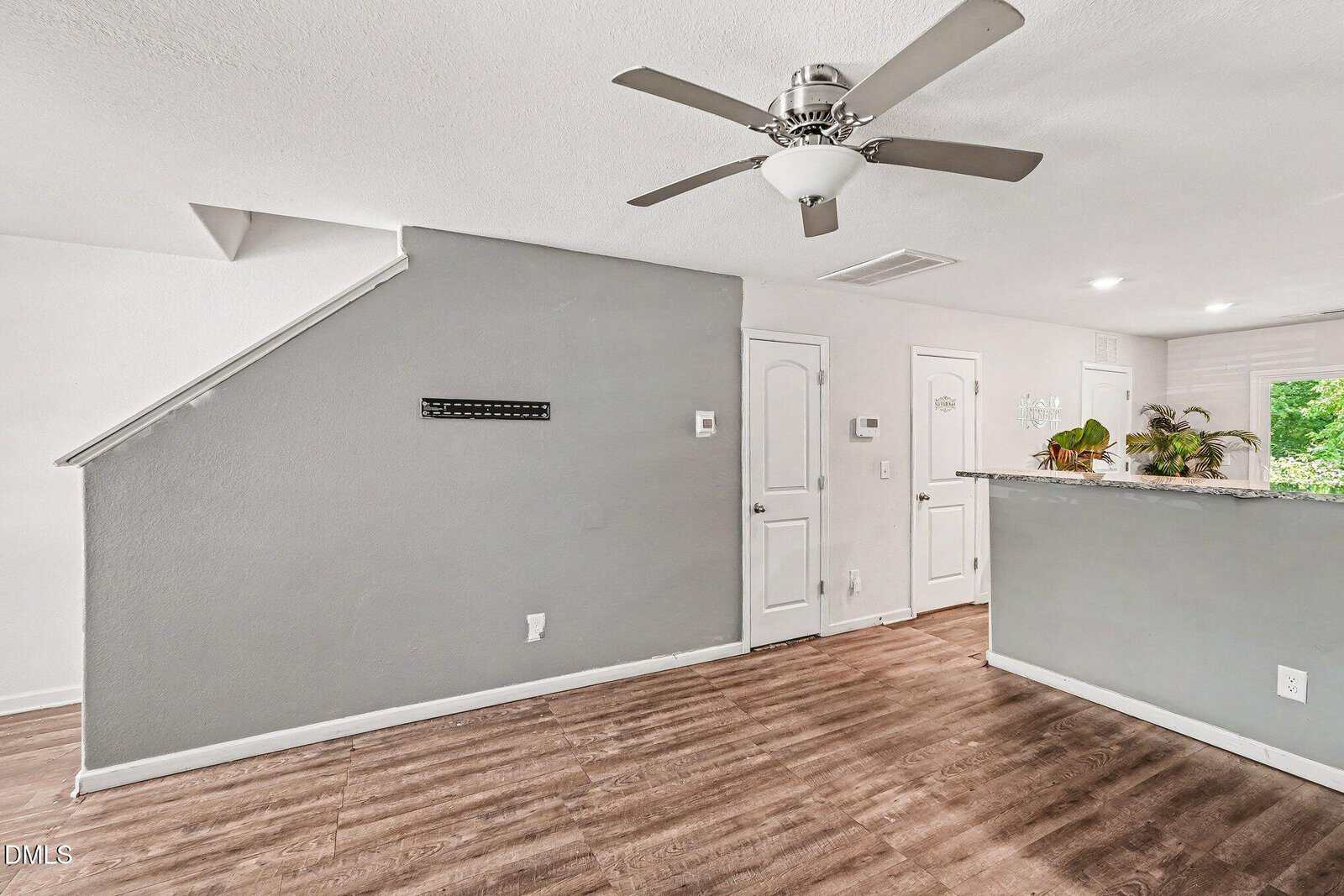 Modern interior with wood flooring, a light gray accent wall under a staircase, ceiling fan, and a kitchen area with gray breakfast bar, white cabinets, and plants on the counter. White doors and bright lighting are visible.