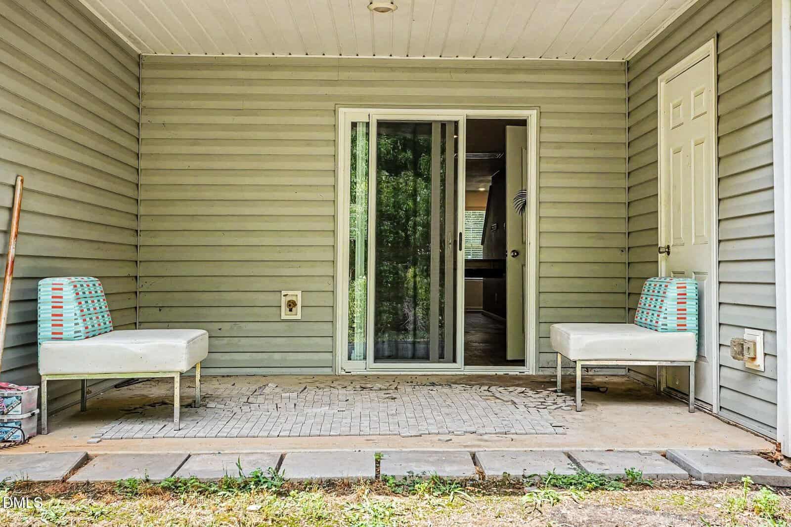 A covered patio with two white chairs featuring blue and orange cushions, a sliding glass door in the center, a closed white door on the right, and light gray siding on the walls. The patio floor is worn and cracked.