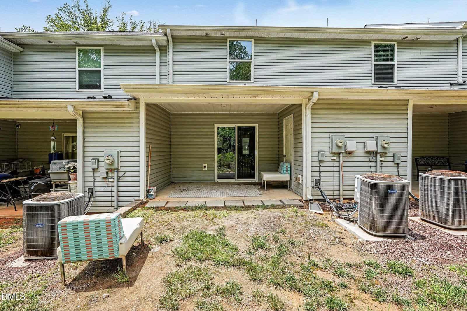 Back view of a two-story townhouse with light green siding, two patio chairs, a small table, sliding glass door, and two outdoor HVAC units on a patchy yard with pavers leading to the entrance.