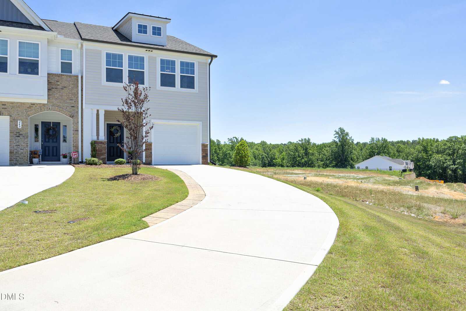 A modern townhouse with brick and light siding, a small tree in front, and a long, curved driveway leading to a garage. The house is next to an open grassy area under a clear blue sky.