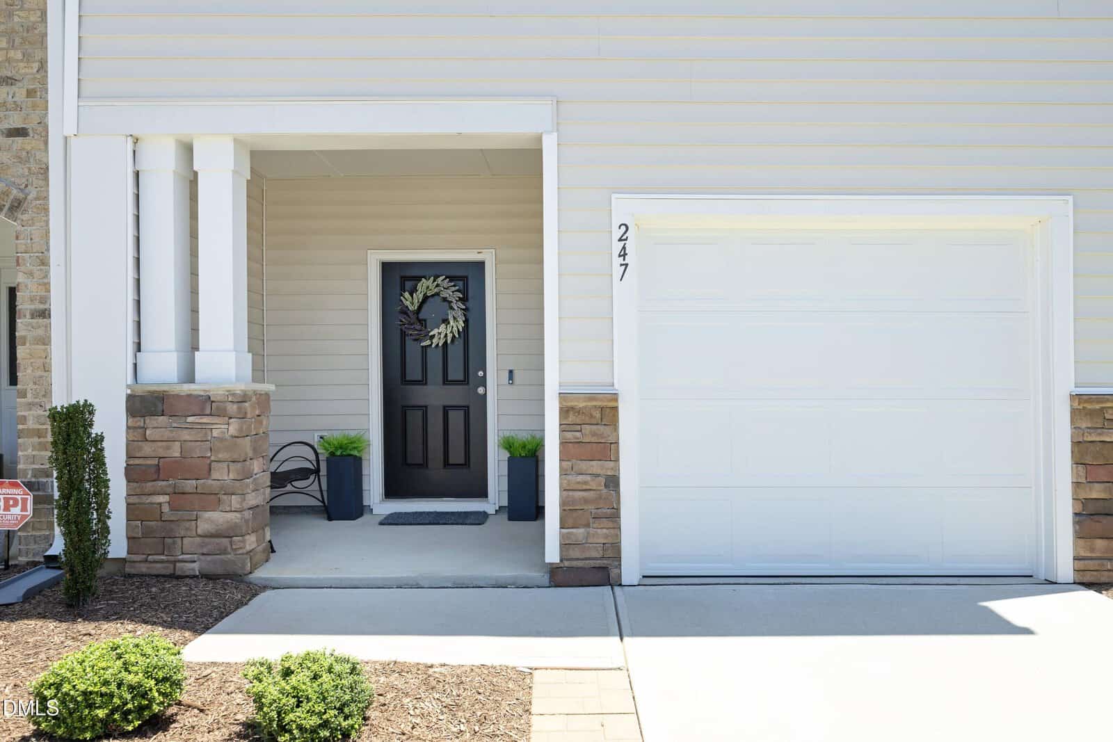A modern house exterior with a black front door decorated with a wreath, two potted plants on either side, a white garage door, and beige siding with brick accents. House number 247 is visible next to the garage.