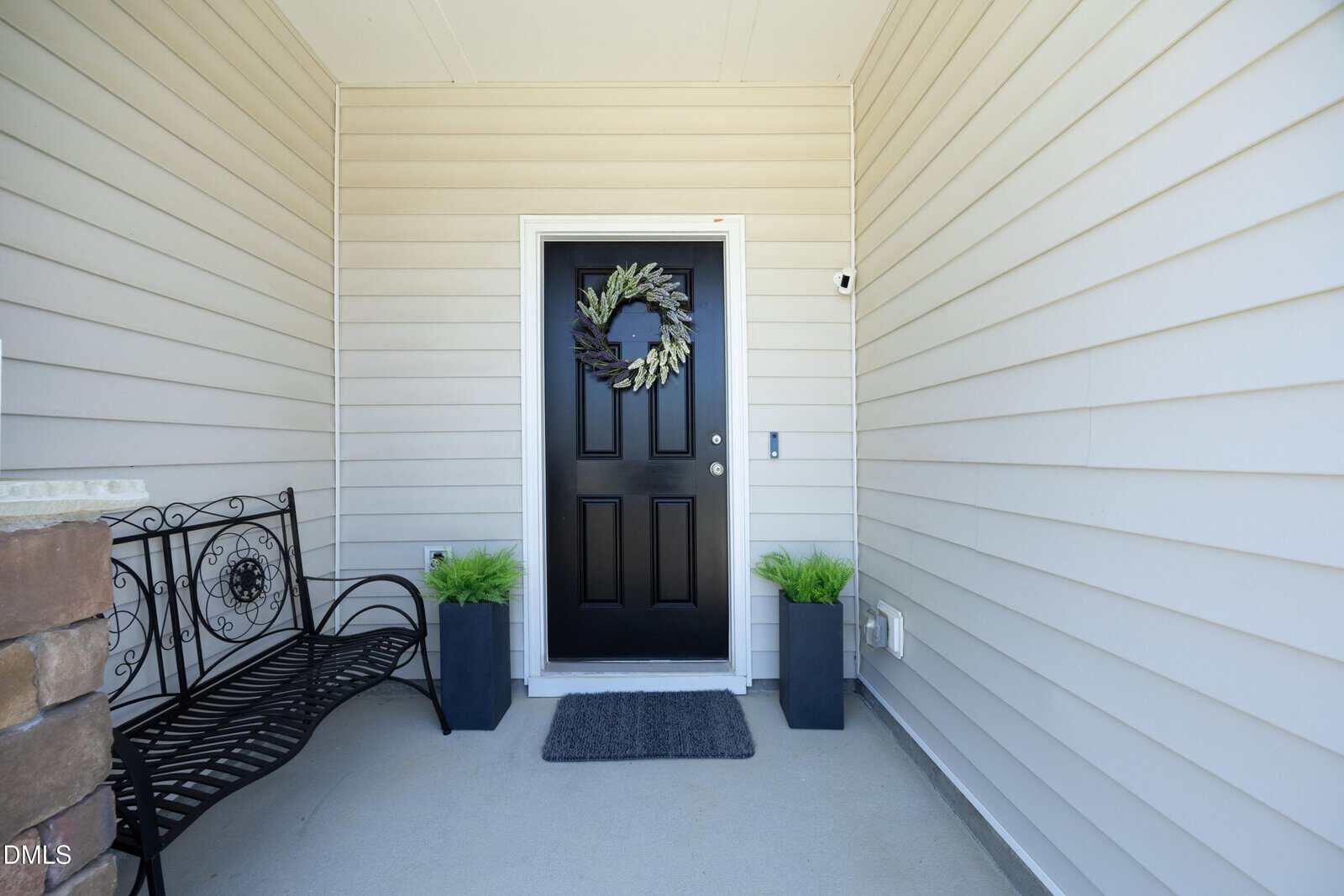 A black front door with a wreath, flanked by two potted plants, sits between beige siding walls. A black bench is on the left, and a gray doormat lies in front of the door.