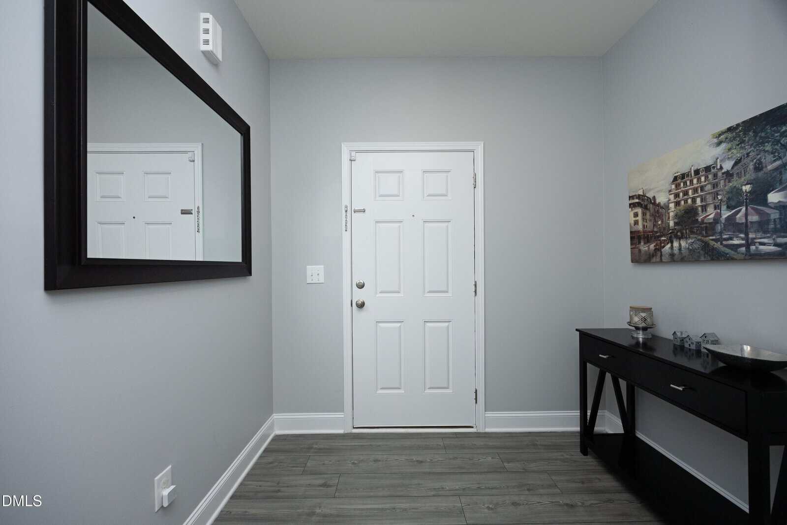 A small, modern entryway with a white door, gray walls, wood-look flooring, a black console table with decor, a large wall mirror, and a painting on the wall.
