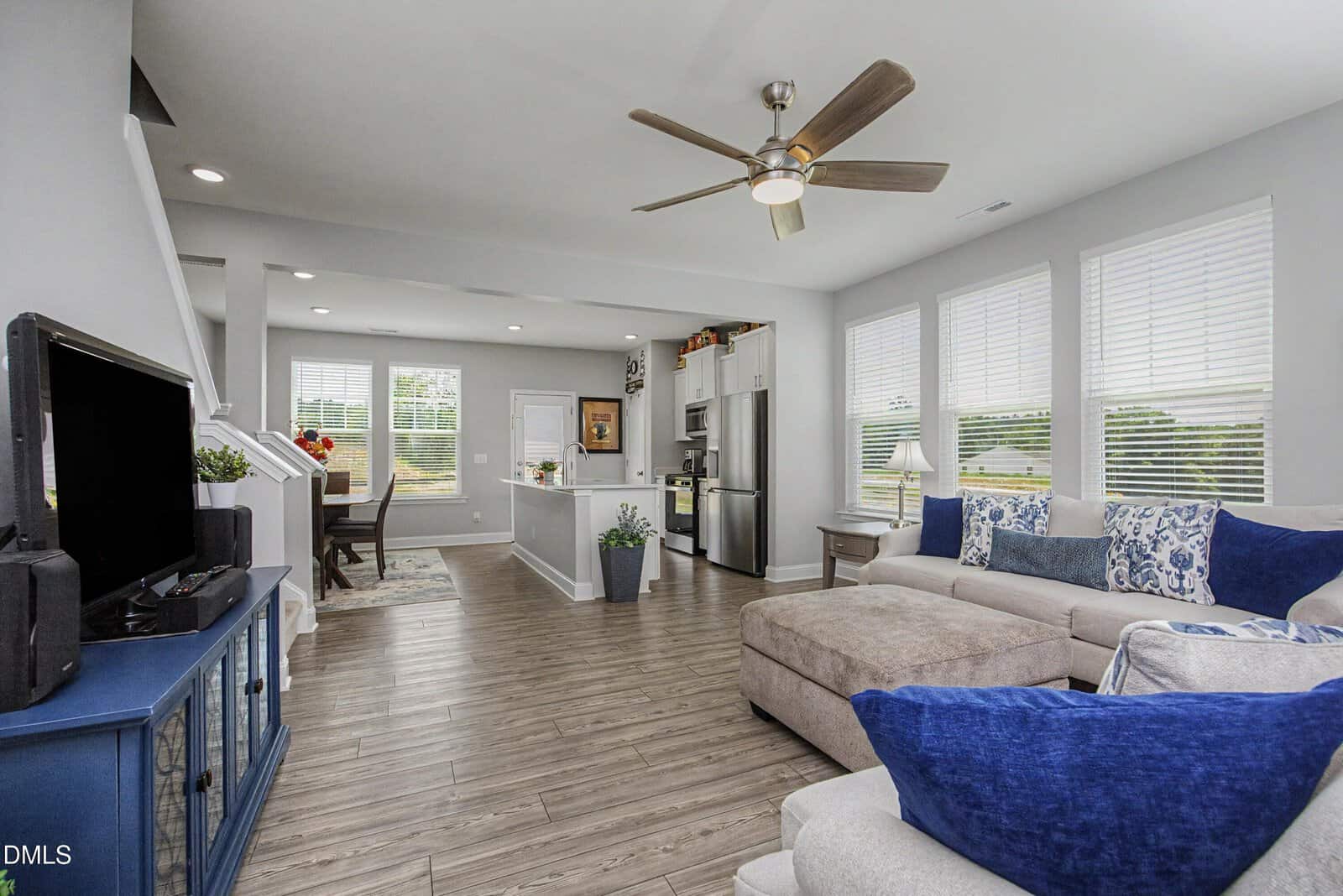 Modern open-concept living room and kitchen with light wood floors, a beige sectional sofa with blue pillows, large windows, a ceiling fan, and a TV on a blue console. Dining area is visible in the background.