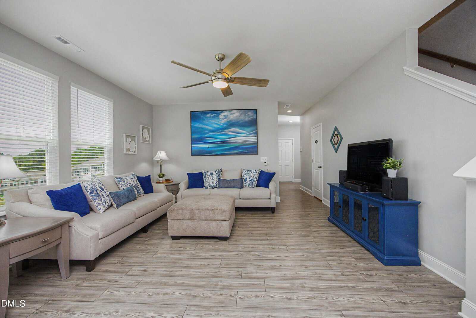 A bright living room with light wood flooring, two beige sofas with blue and white cushions, a central ottoman, a blue console under a TV, wall art, and large windows bringing in natural light.