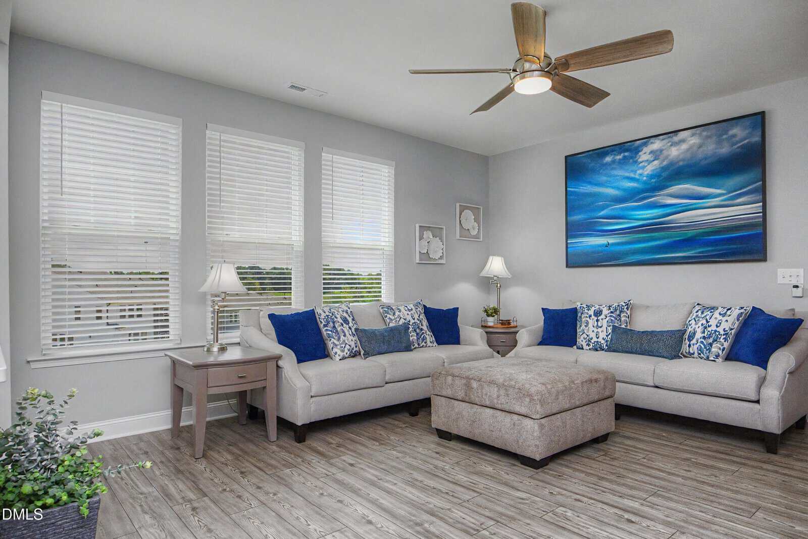 Modern living room with two light gray sofas, blue and white pillows, a matching ottoman, wooden floor, large artwork on the wall, and windows with white blinds letting in natural light. A ceiling fan hangs above.