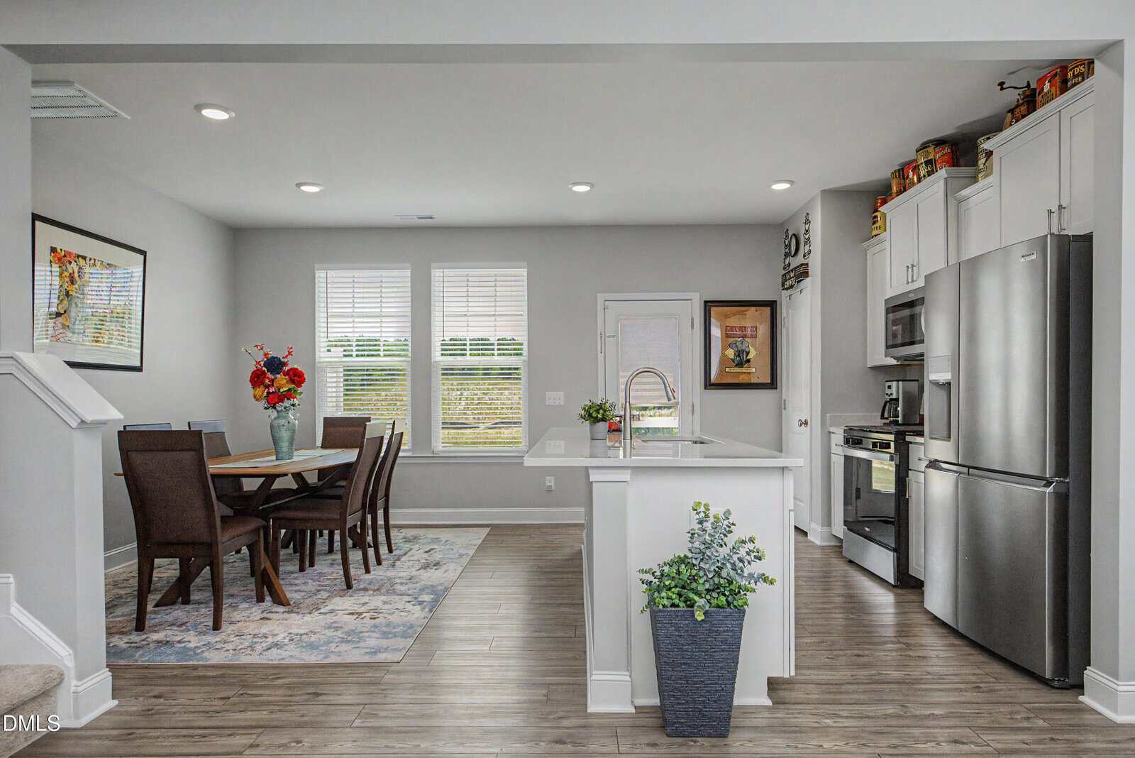 Modern kitchen and dining area with white cabinets, stainless steel appliances, a dining table with six chairs, colorful flowers on the table, and decorative plants, all in a bright space with wood flooring and large windows.