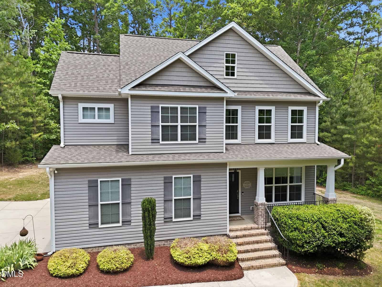Two-story gray house with white trim, front porch, and black door at 3689 Summer Springs Drive in Franklinton. Well-maintained landscaping features bushes, mulch, a tall shrub by the entrance, plus a driveway on the left surrounded by trees.