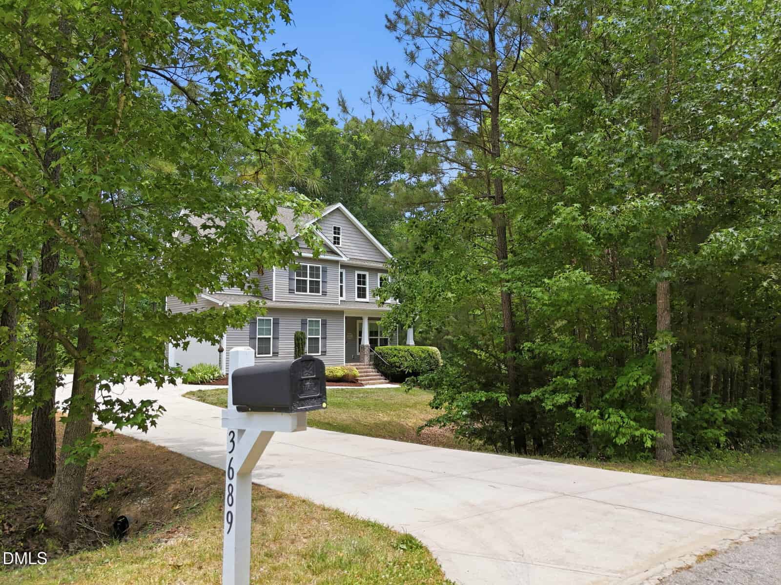 A two-story gray house with white trim sits behind a long driveway, surrounded by tall green trees. A black mailbox with the number 36809 stands at the curb near the driveway entrance.