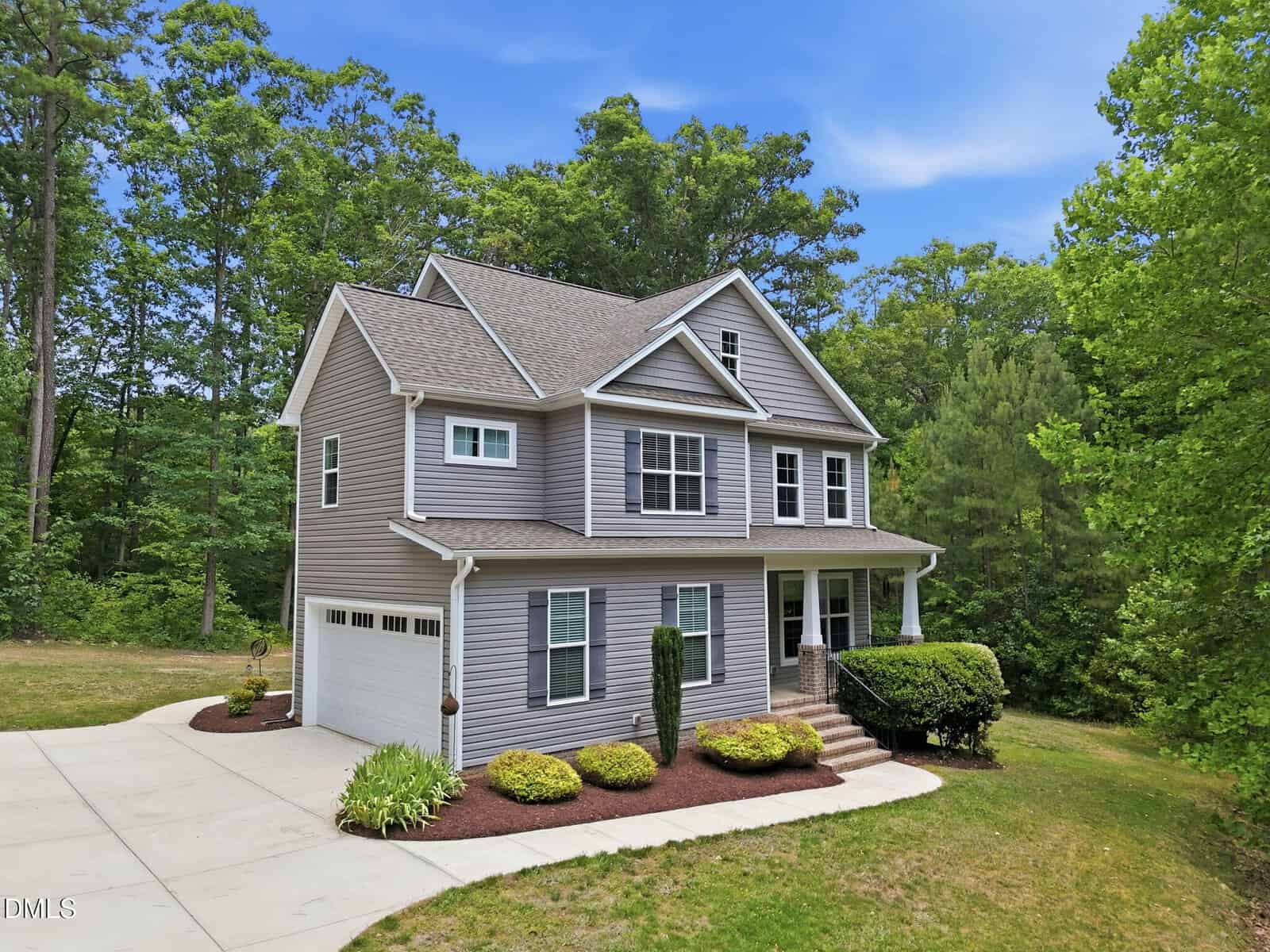 A two-story gray house with white trim, a front porch, and attached garage sits on a manicured lawn with shrubs, surrounded by tall trees and greenery under a blue sky.