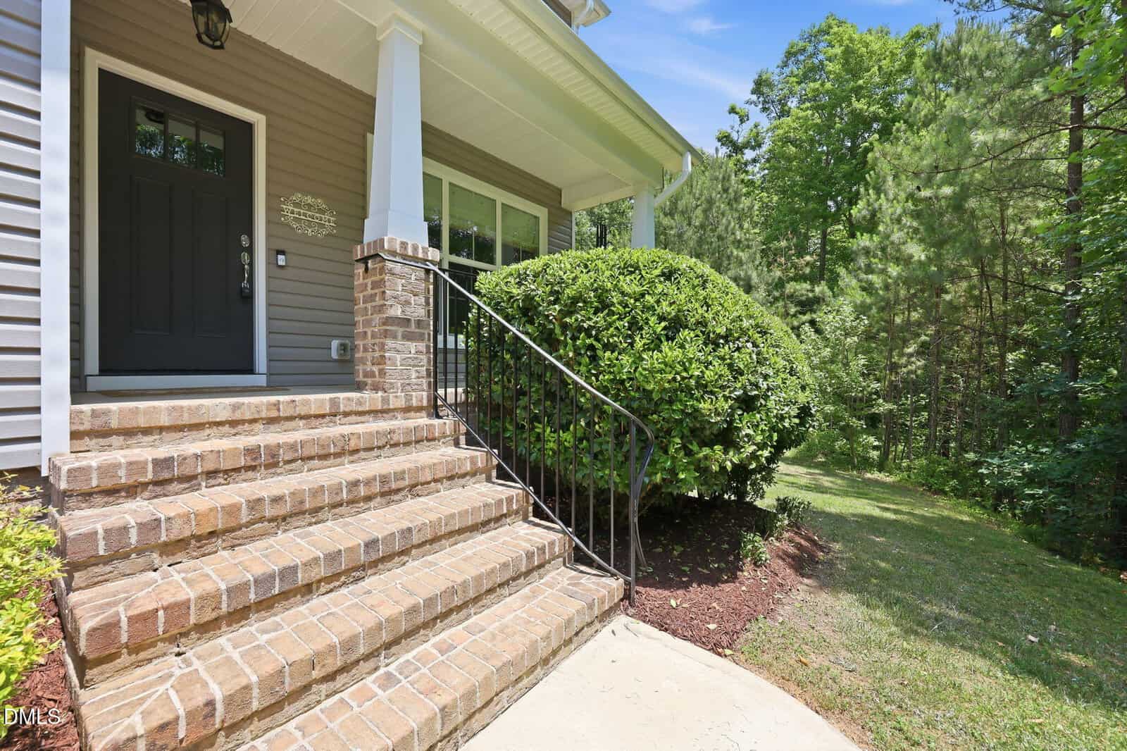 Brick steps with a black metal railing lead up to a gray front door of a house with white trim. Large windows and trimmed bushes are by the entrance, with a grassy yard and tall trees in the background.