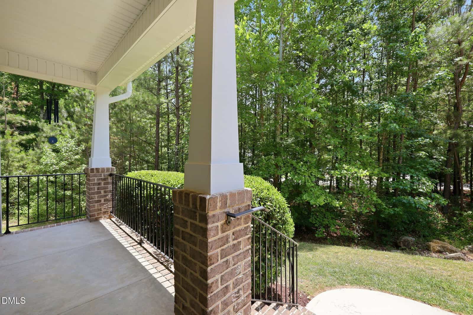 View from a covered porch with white columns and brick bases, black metal railing, and a concrete floor, overlooking a lush green yard bordered by trees and shrubs on a sunny day.