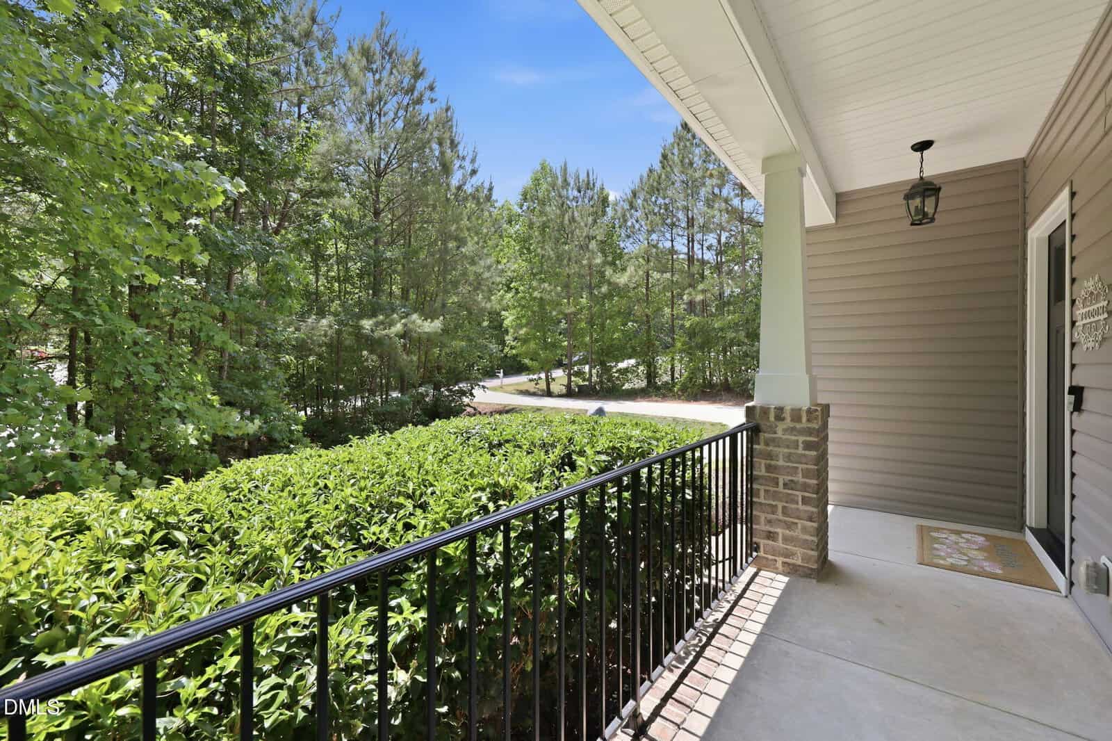 A covered front porch with a black metal railing, brick column, beige siding, and a view of green bushes, trees, and a quiet road under a bright blue sky.