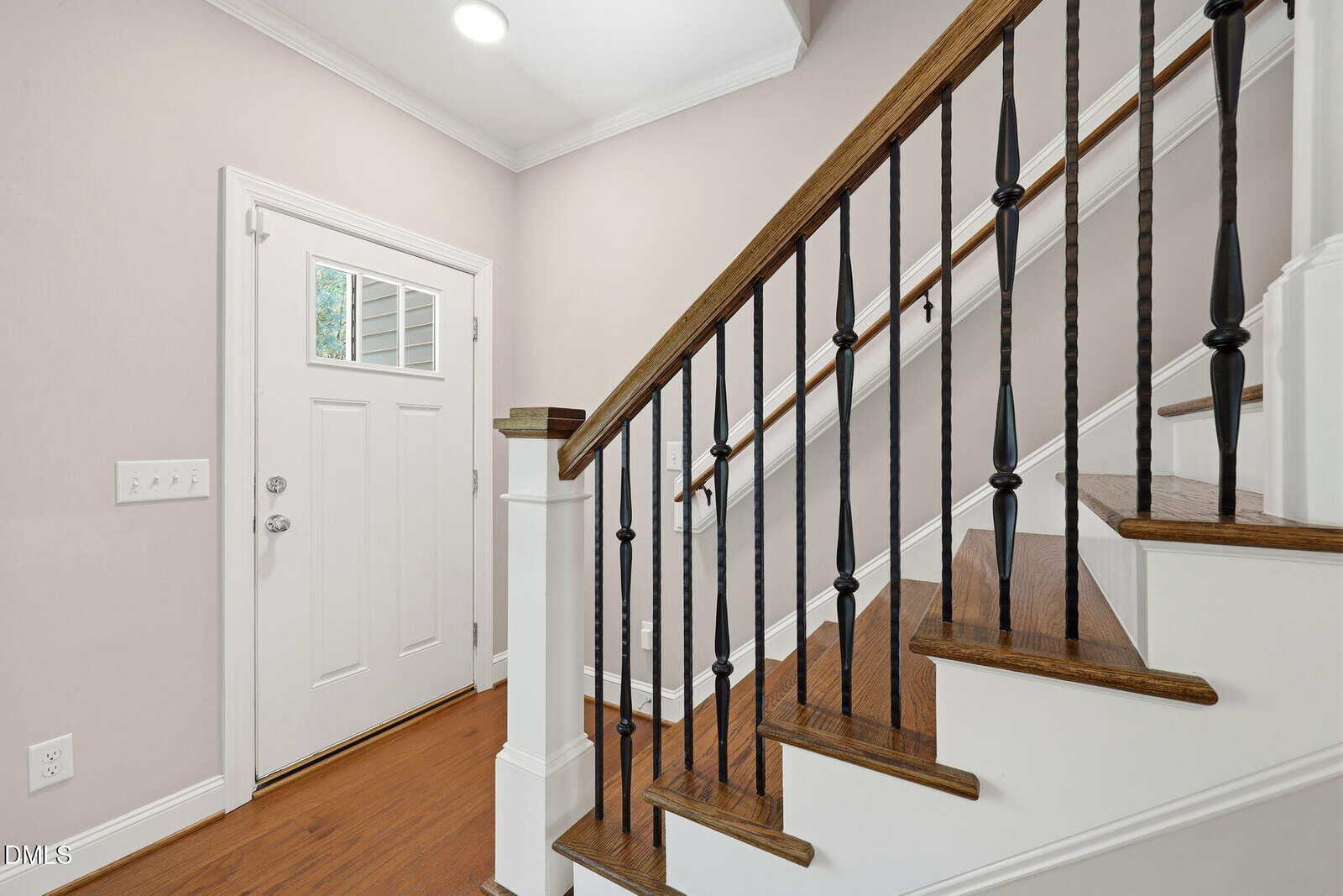 A bright entryway with a white door, hardwood floors, and a wooden staircase with white risers and black metal balusters.