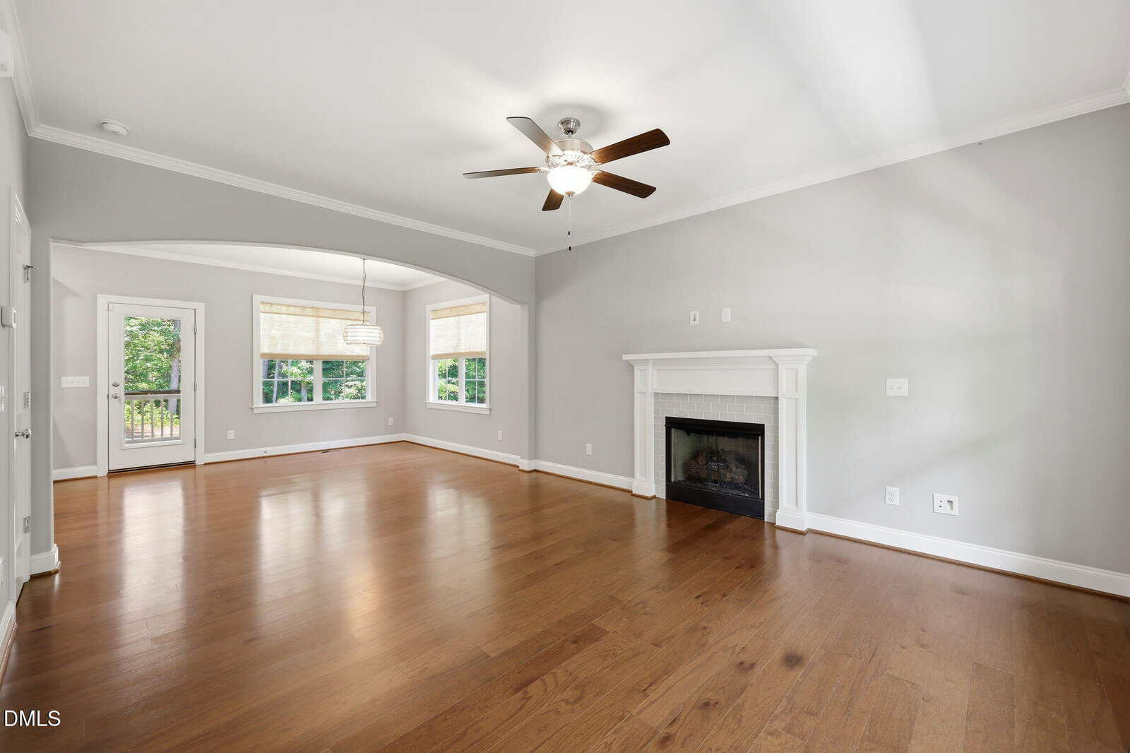 Spacious, empty living room with hardwood floors, a ceiling fan, fireplace, and large windows letting in natural light. There’s an open archway leading to a dining area with more windows and a glass door to the outside.