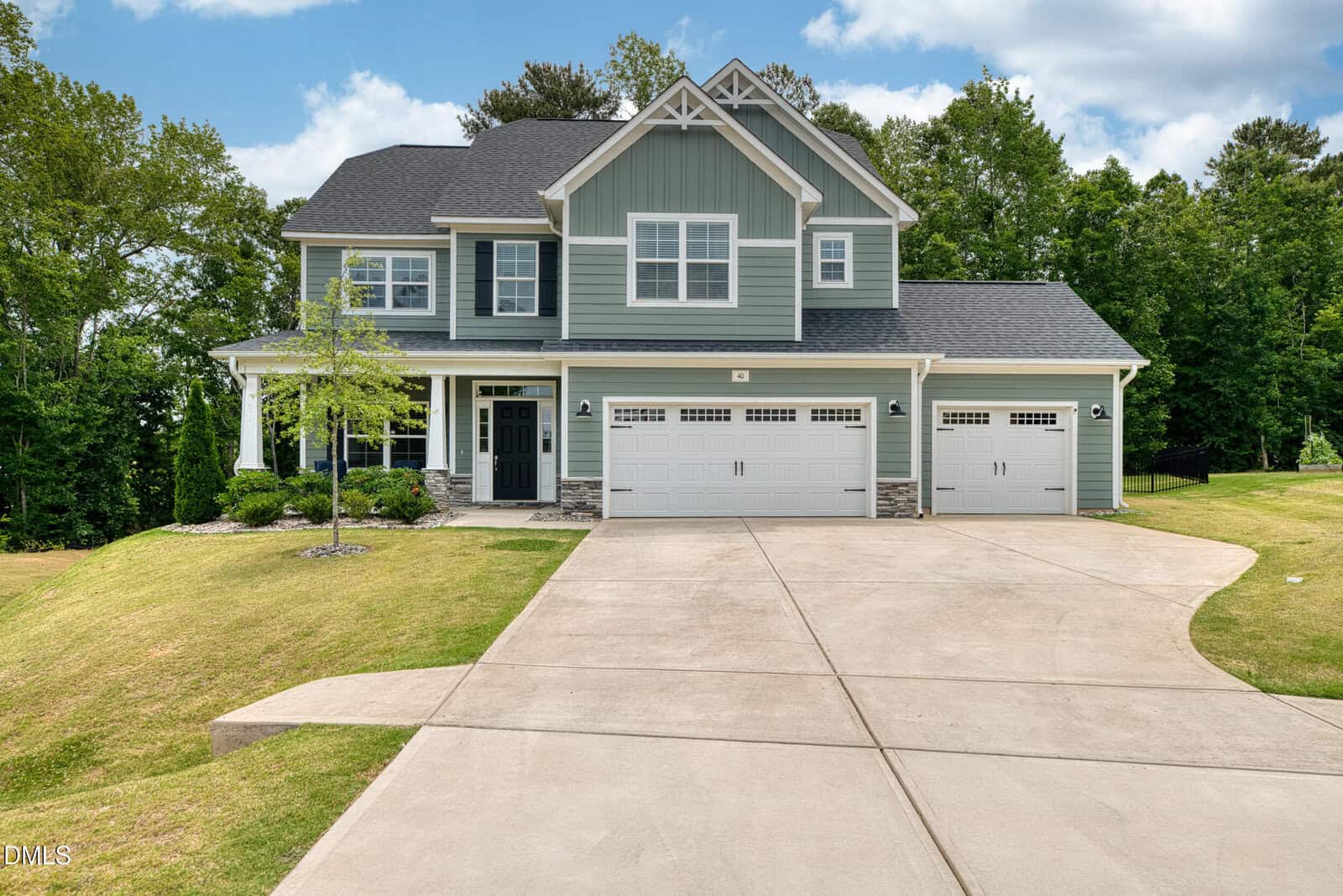 A two-story green house with white trim at 40 Cowpens Court in Franklinton, featuring a covered front porch, three-car garage, and large driveway, surrounded by a well-kept lawn and trees under a blue sky with clouds.