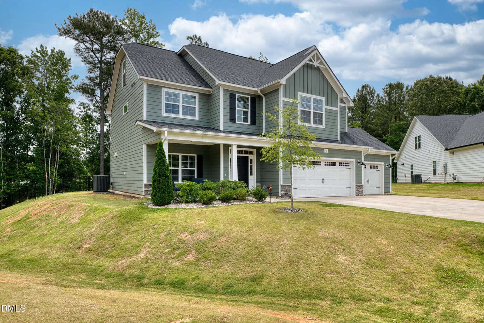 Two-story green house with white trim, front porch, and attached double garage, set on a grassy, slightly sloped lawn with small shrubs, a young tree, and a concrete driveway on a sunny day.