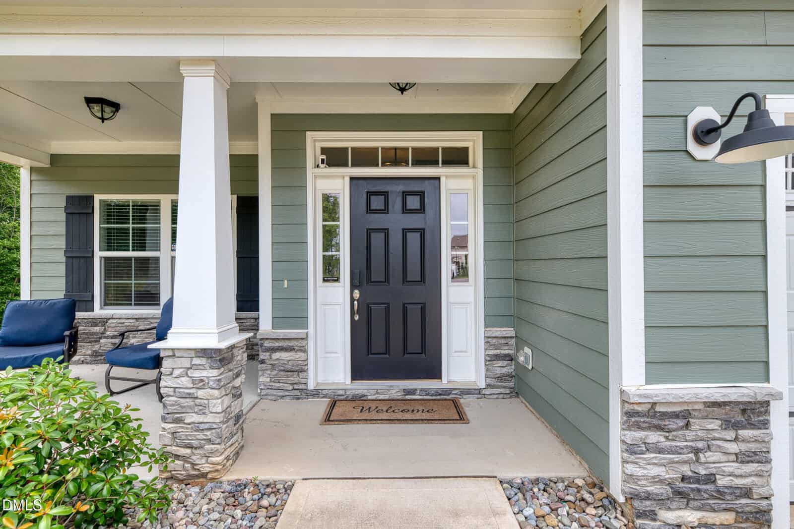Front porch of a house with green siding, stone accents, white trim, and a black front door. A "Welcome" doormat sits at the entrance, and two blue cushioned chairs are visible on the left side.