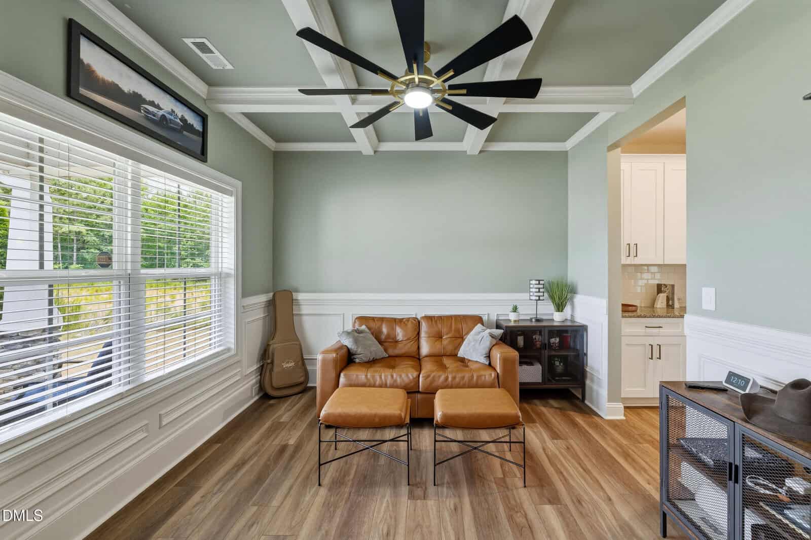 A cozy living room with a brown leather loveseat and two ottomans, a guitar in the corner, wood flooring, large windows, ceiling fan, and mint green walls. A kitchen area is visible through an open doorway.
