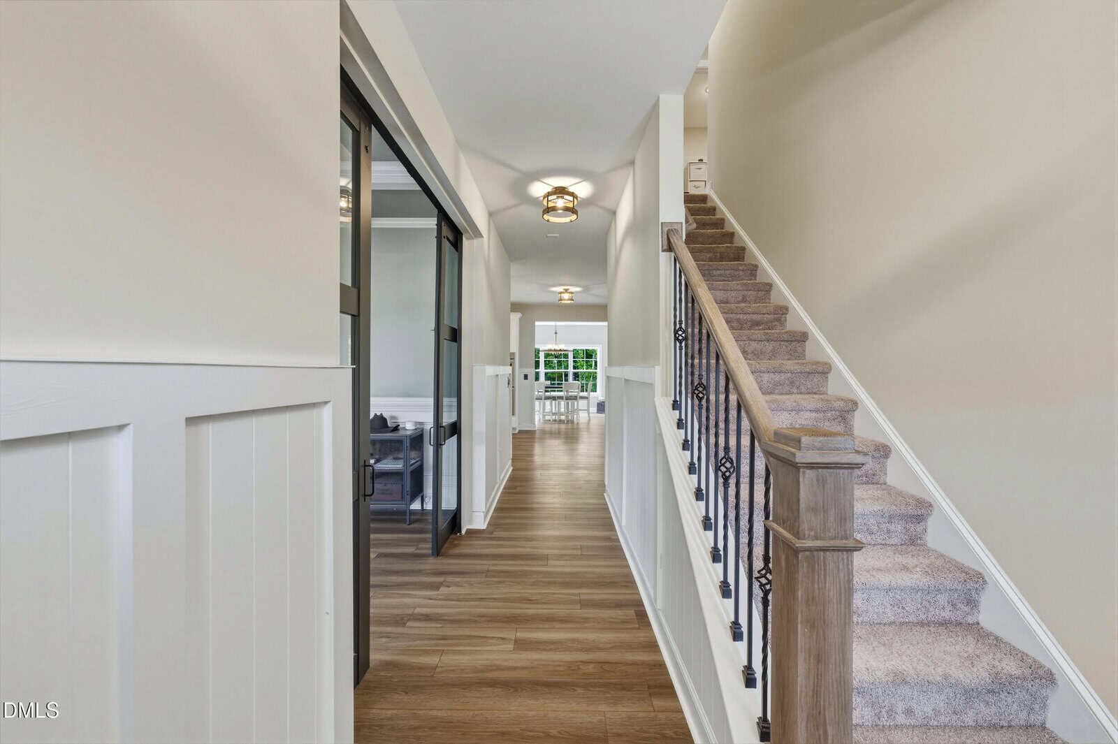 A hallway with wood flooring and white walls leads to a bright area. Carpeted stairs with a wooden banister are on the right, and a glass-paneled room is on the left. Ceiling lights illuminate the space.