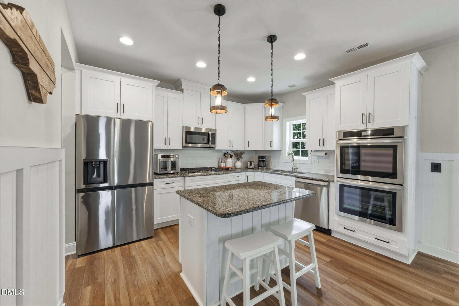 Modern kitchen with white cabinets, stainless steel appliances, a center island with granite countertop, two white barstools, pendant lights, wooden floors, and bright lighting.