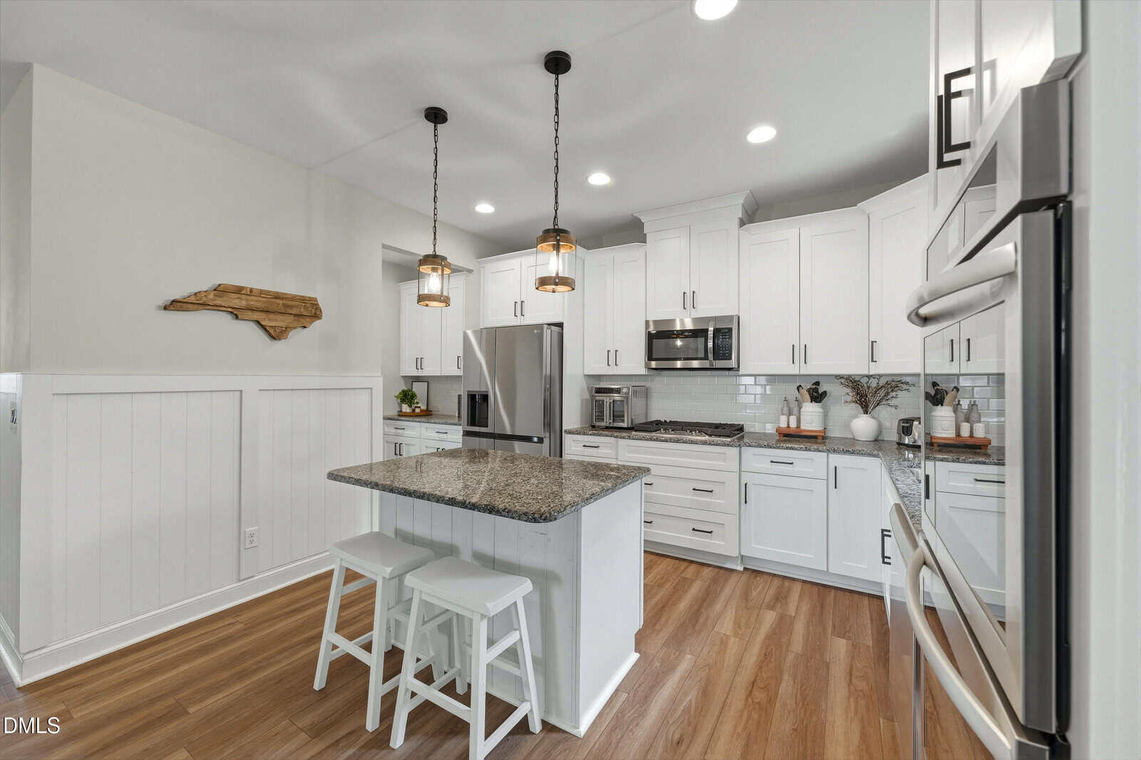 Modern kitchen with white cabinets, stainless steel appliances, granite island with two white stools, wood floors, pendant lights, and a wood wall art piece on a white paneled wall.