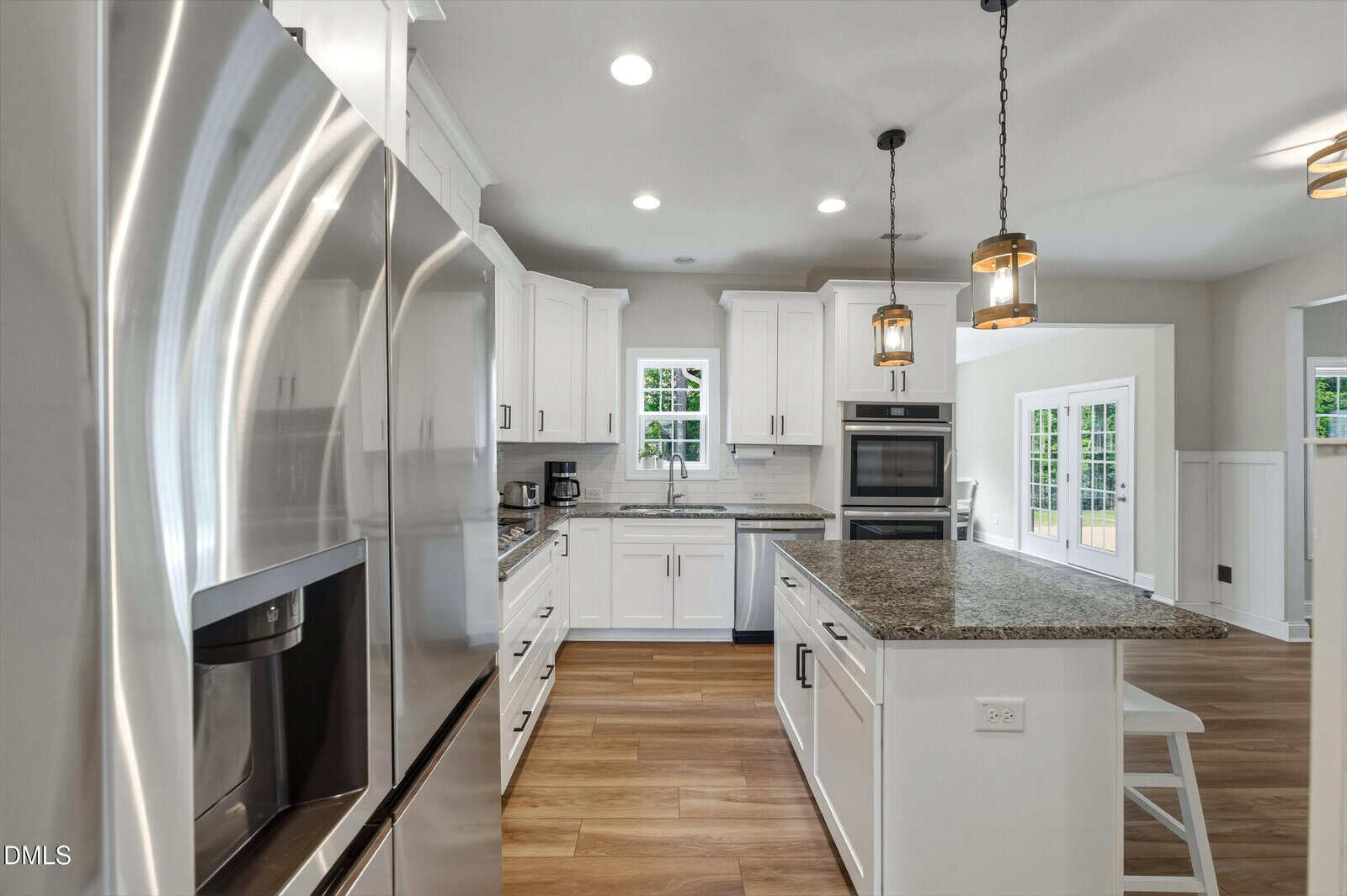 Modern kitchen with white cabinets, stainless steel appliances, granite countertops, a center island with stools, pendant lights, hardwood floors, and large windows letting in natural light.