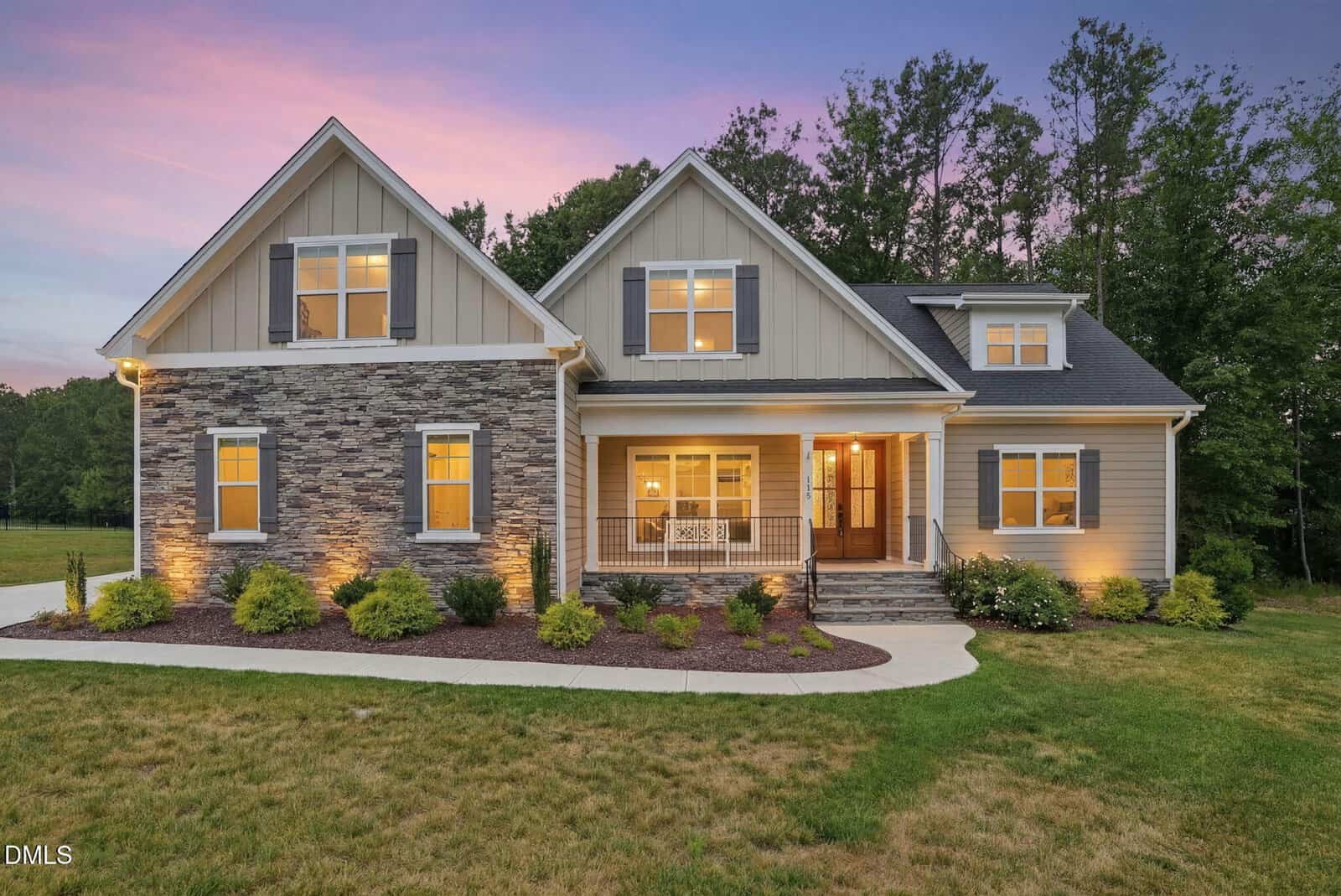 A modern suburban house at 115 Old Garden Lane, Youngsville, features a mixed stone and siding exterior, warmly lit windows, a covered porch, and landscaped front yard set against a backdrop of trees at sunset.