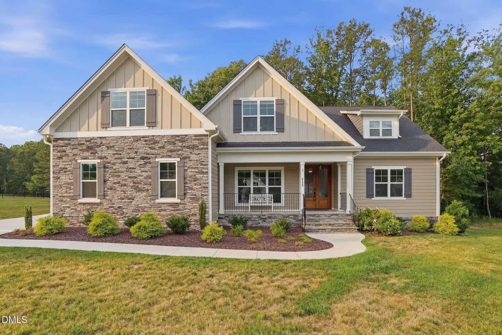 A modern suburban house at 115 Old Garden Lane, Youngsville, featuring tan siding, stone accents, a front porch with a wooden door, manicured shrubs, and a green lawn, set against a backdrop of trees and blue sky.
