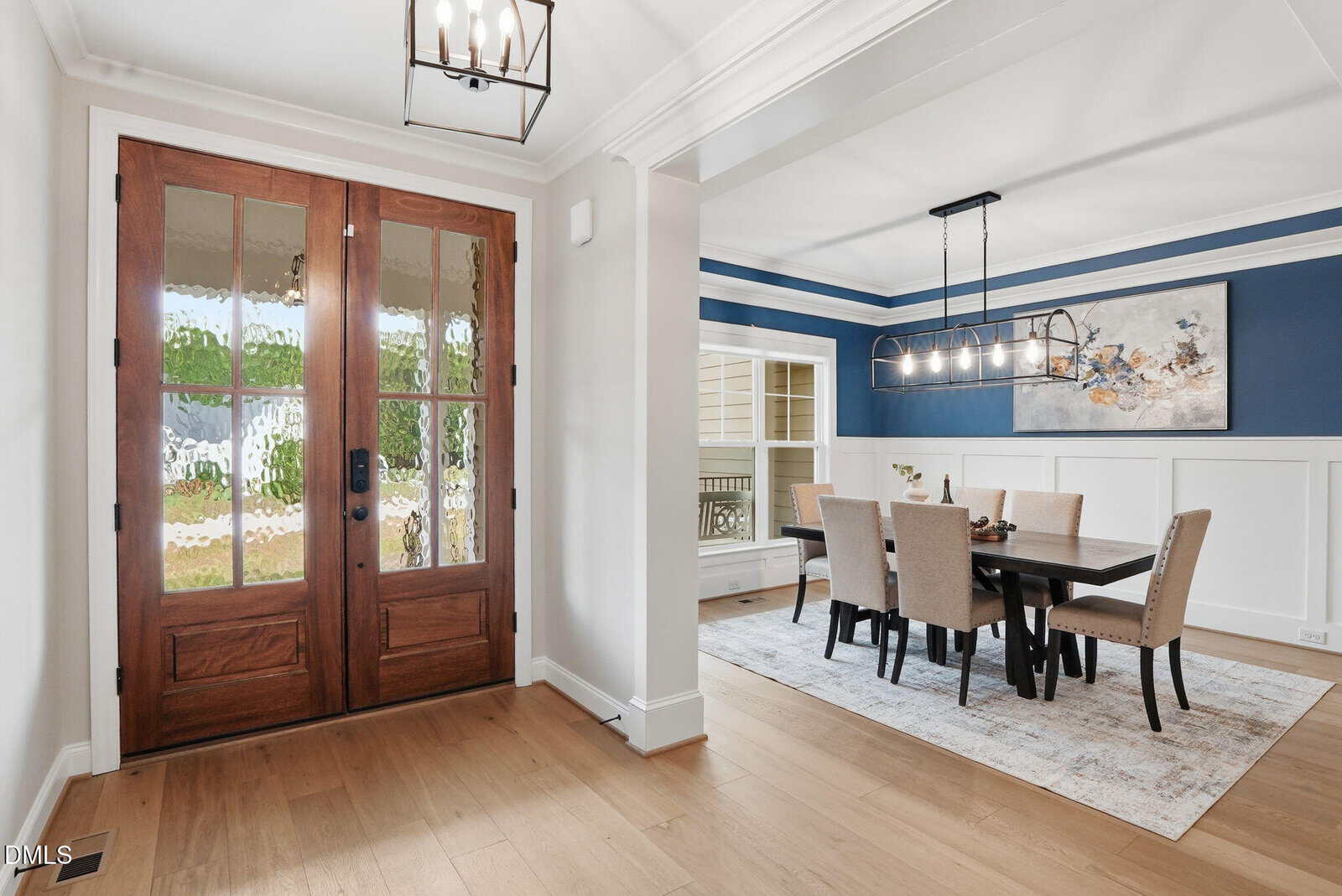 A bright entryway with wooden double doors leads to a dining room featuring a black table, eight beige chairs, a modern chandelier, blue accent wall with wainscoting, and a large abstract painting.