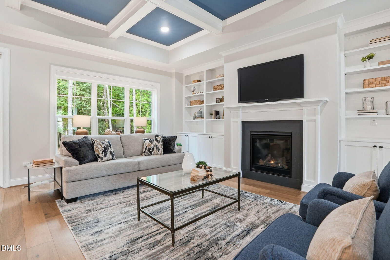 Modern living room with a gray sofa, glass coffee table, two blue chairs, built-in shelves, a fireplace, wall-mounted TV, and large window showing green trees outside. Room features wood floors and a coffered, blue-accented ceiling.