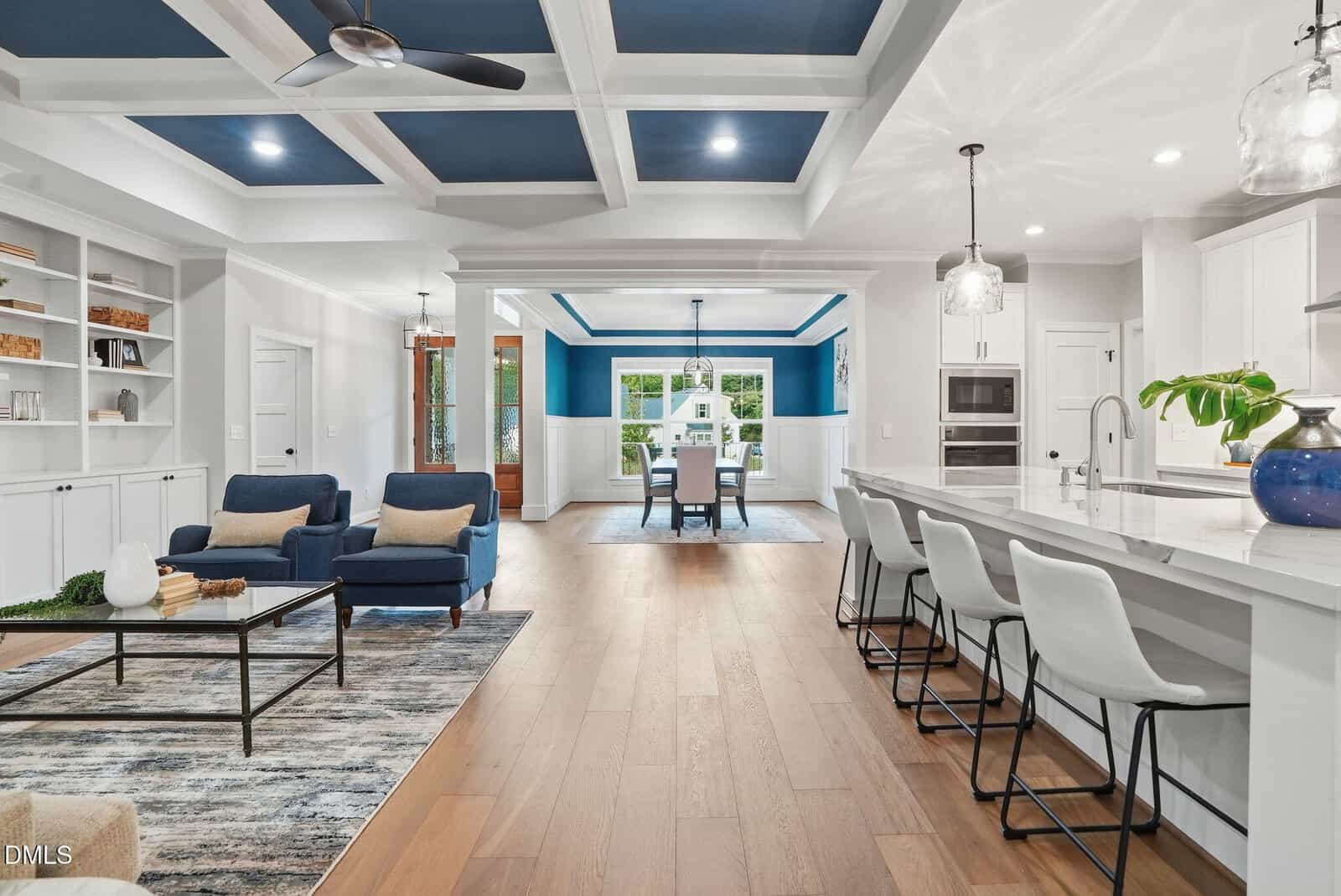 Spacious open-concept living area with blue and white accents, modern kitchen with island seating, dining area in the background, and built-in shelves on the left.