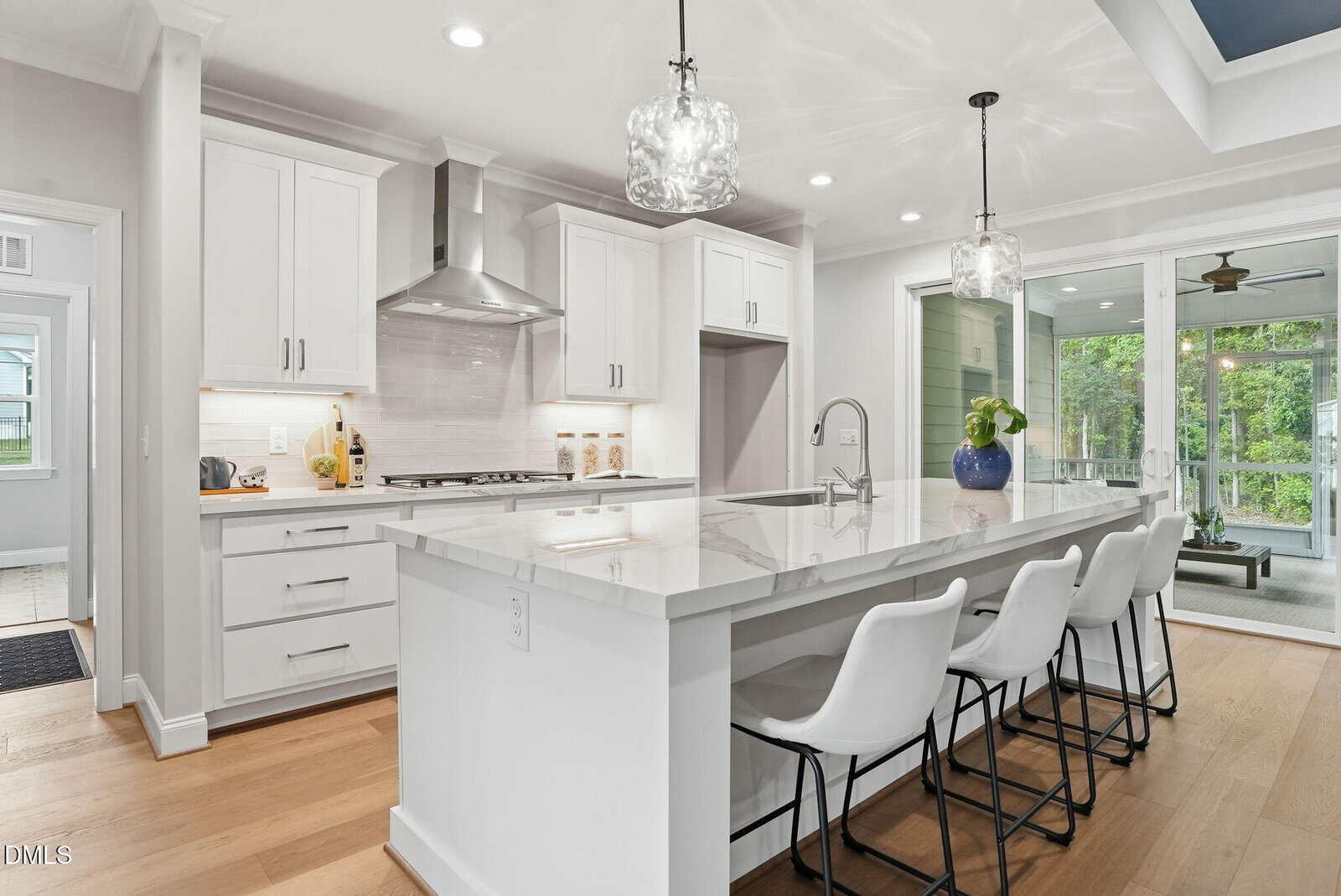 Modern white kitchen with a large marble island, four white barstools, stainless steel appliances, pendant lights, and glass doors opening to a screened porch with green trees visible outside.
