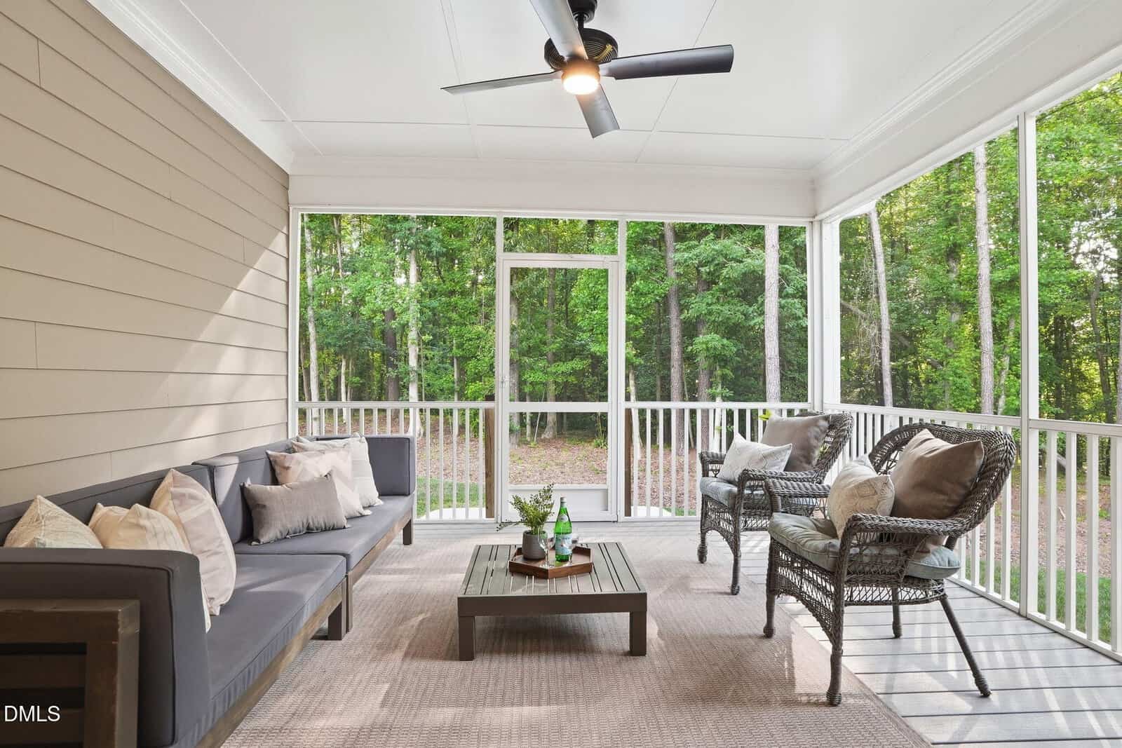 A screened-in porch with a ceiling fan, outdoor sofa, two wicker chairs with cushions, a low coffee table, and forest views through the screened walls. The decor is neutral and inviting.