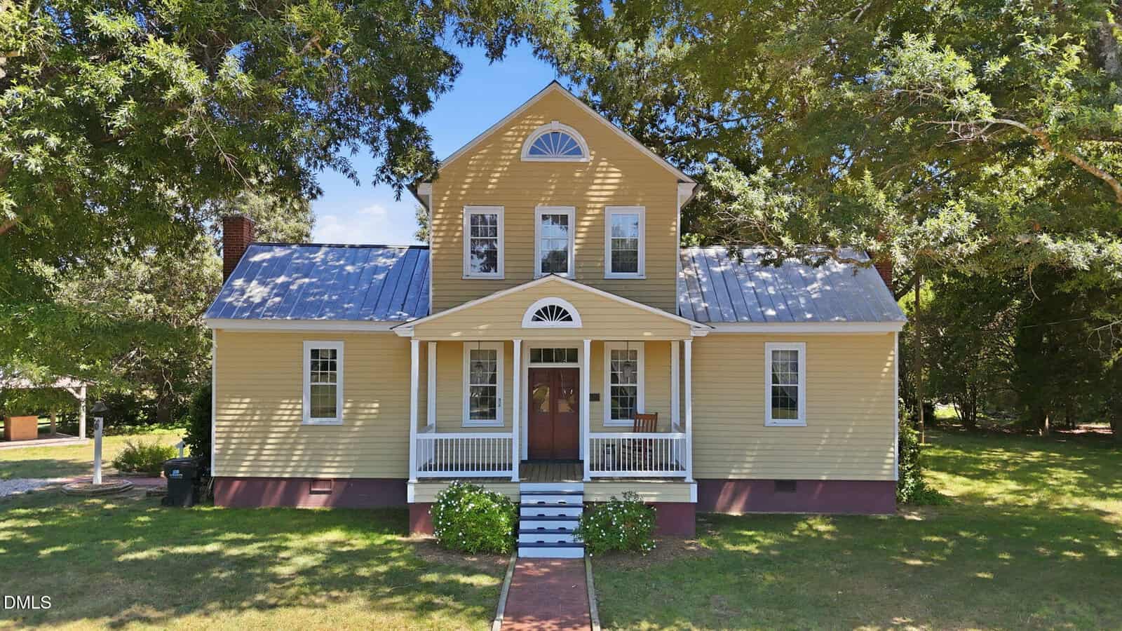 A yellow two-story house with a metal roof, white trim, and a small front porch with steps and rails sits at 462 Darius Pearce Road in Youngsville. Trees surround the home, casting partial shade on the lawn and building.