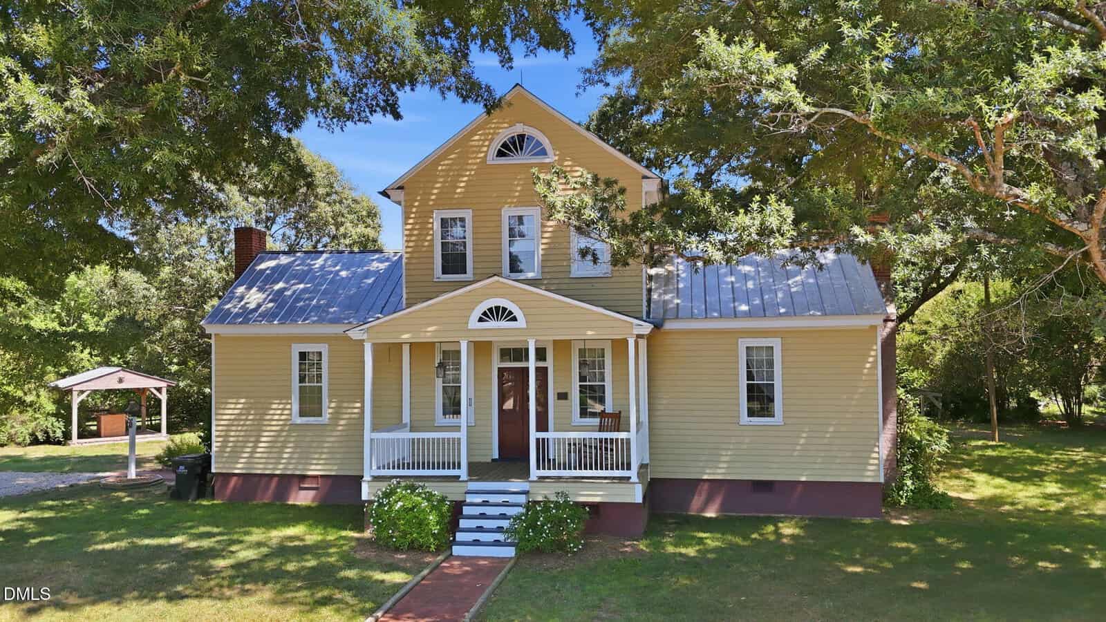 A yellow two-story house with a metal roof, white trim, and a small covered front porch, surrounded by green grass, trees, and a detached carport in the background.