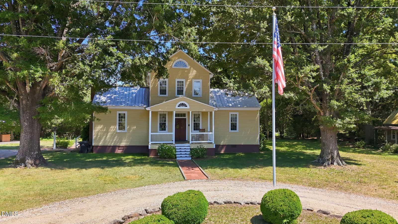 A yellow two-story house with a covered front porch and white trim sits behind a circular gravel driveway, surrounded by trees and neatly trimmed bushes, with an American flag on a pole in the front yard.