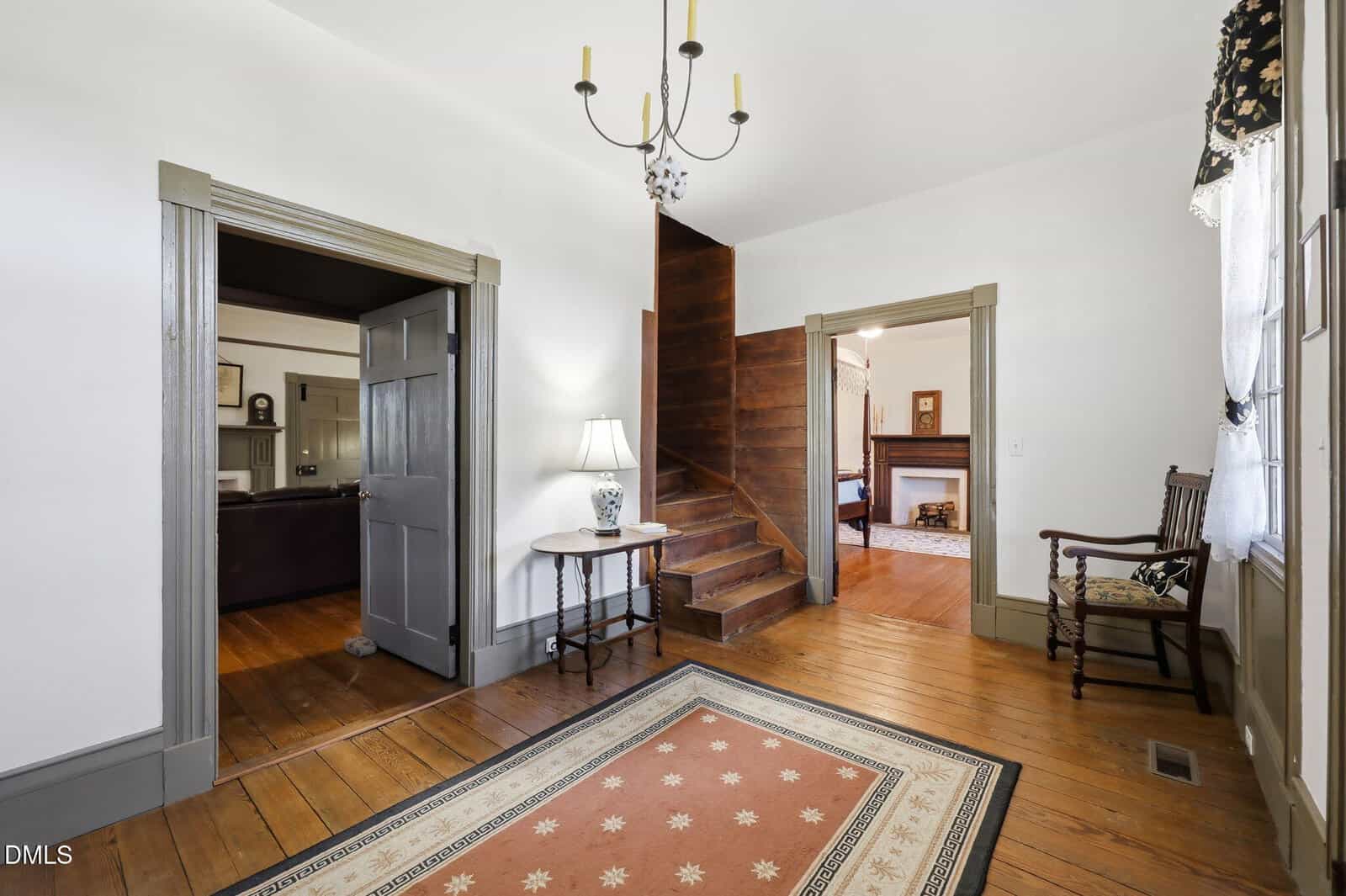 Bright entryway with wood floors, patterned rug, wooden staircase, a round table with a lamp, a wooden chair by a window, and open double doors leading to another room with a fireplace.